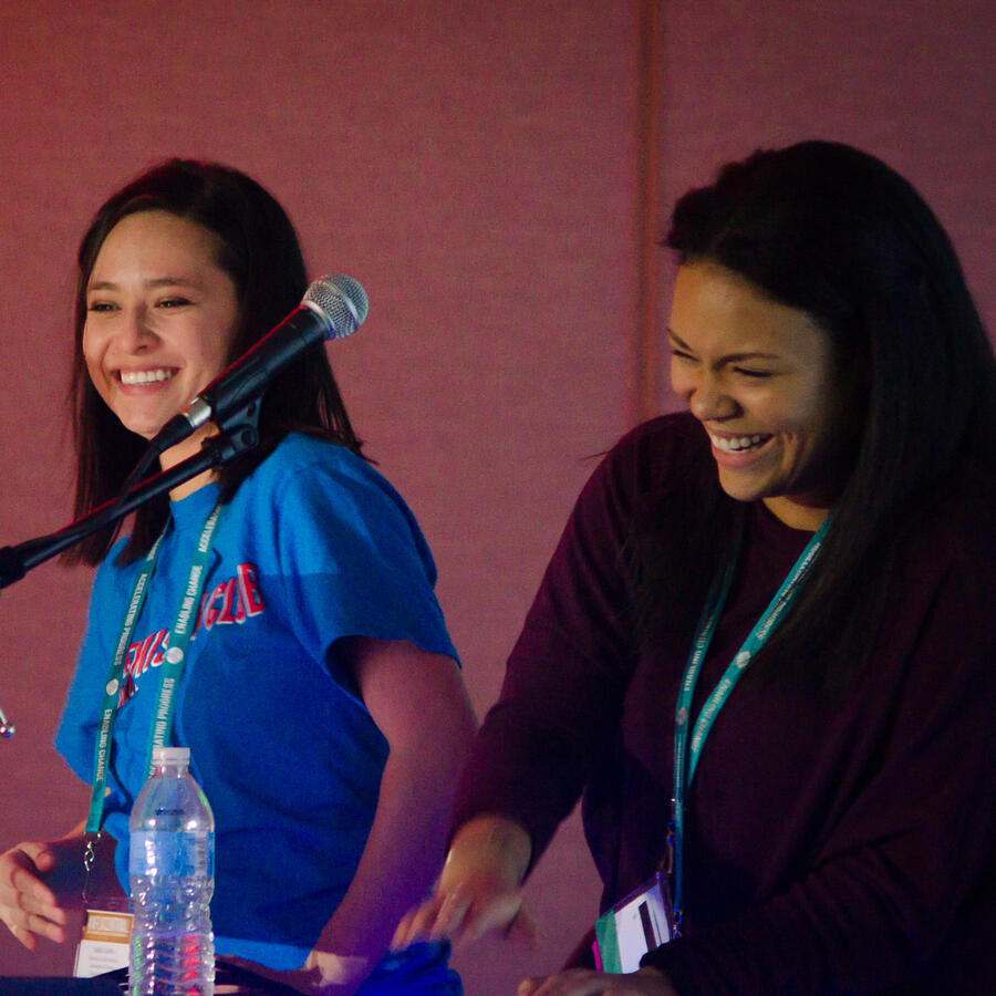 Two women at a podium with microphones laugh under warm stage lighting.