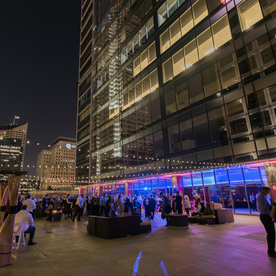 Nighttime crowd mingles on a high-rise patio with city skyline reflections and bistro lights overhead.