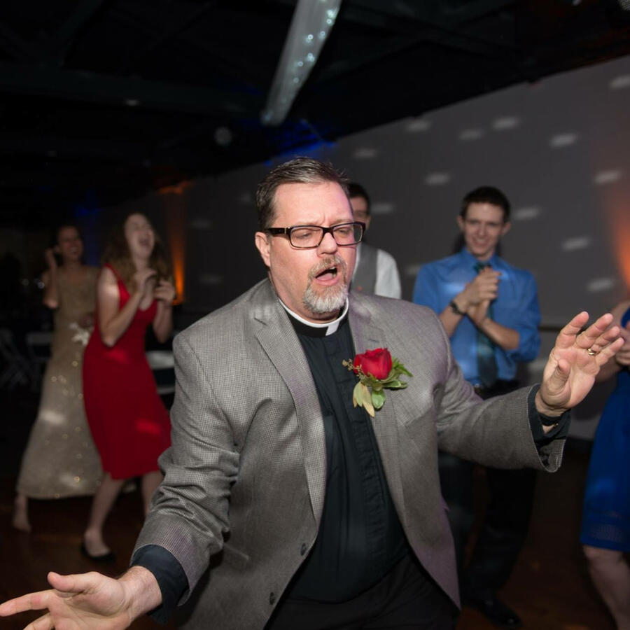 Man in a clerical collar dances on a crowded floor while guests cheer in the background.